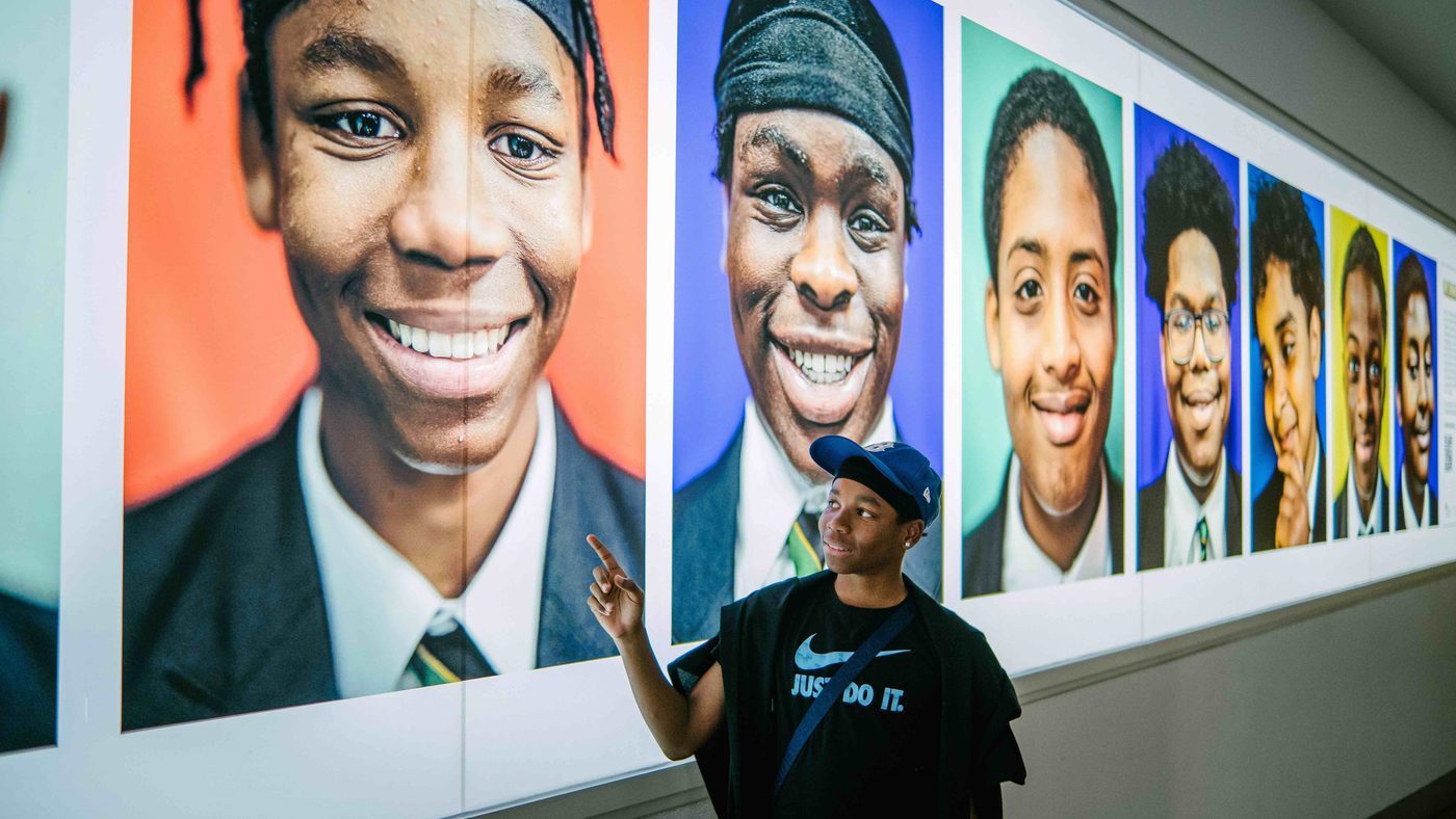 A young man stands by a wall of photos taken by Kay Rufai as part of his Smiling Boys Project. They are all photos of Black school boys smiling.