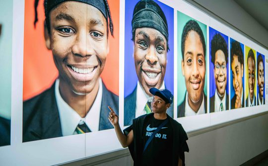 A young man stands by a wall of photos taken by Kay Rufai as part of his Smiling Boys Project. They are all photos of Black school boys smiling.
