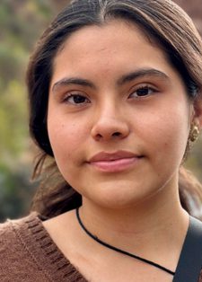 A woman with a soft expression stands outdoors, wearing a brown sweater.