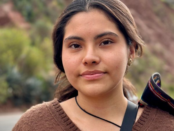 A woman with a soft expression stands outdoors, wearing a brown sweater.