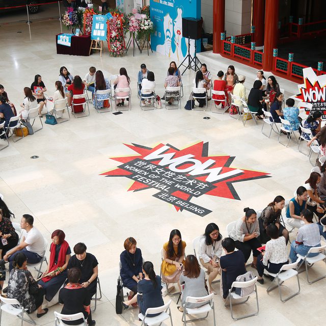 A diverse group of people sits in two large circles of chairs on a tiled floor. The centre features a "Wow" graphic in bold colours