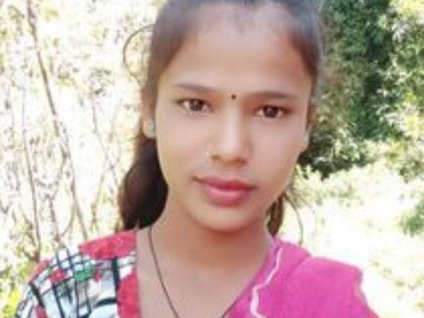 A young woman stands outdoors, wearing a colorful patterned top and a pink scarf