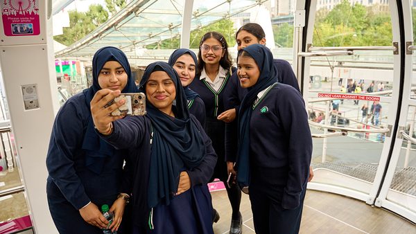A group of six smiling young women in school uniforms take a selfie inside a glass observation pod, with greenery visible outside