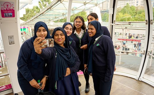 A group of six smiling young women in school uniforms take a selfie inside a glass observation pod, with greenery visible outside