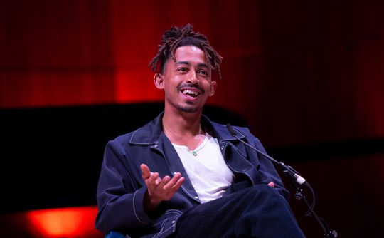 Singer Jordan Stephens sits onstage, gesturing with one hand. He wears a dark jacket. Background is softly lit with warm red tones.