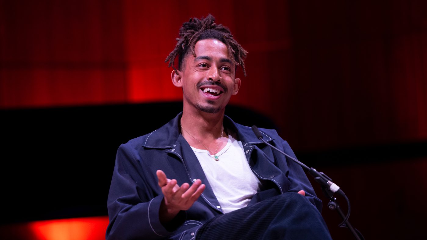 Singer Jordan Stephens sits onstage, gesturing with one hand. He wears a dark jacket. Background is softly lit with warm red tones.
