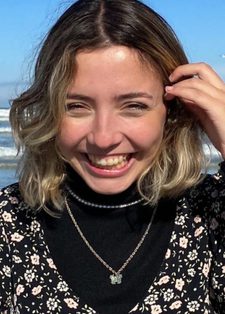 Young woman with a beaming smile stands on a beach. She wears a floral dress over a black turtleneck