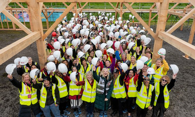 A large group of people in yellow vests and hard hats cheerily pose under a timber structure.