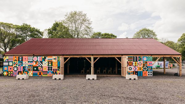 A vibrant wooden barn with a red roof and colourful geometric patterns on the walls, surround by trees and a cloudy sky