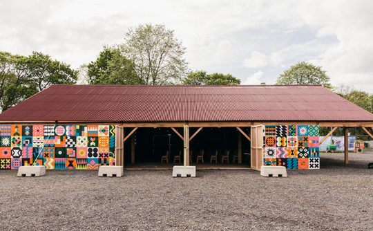 A vibrant wooden barn with a red roof and colourful geometric patterns on the walls, surround by trees and a cloudy sky