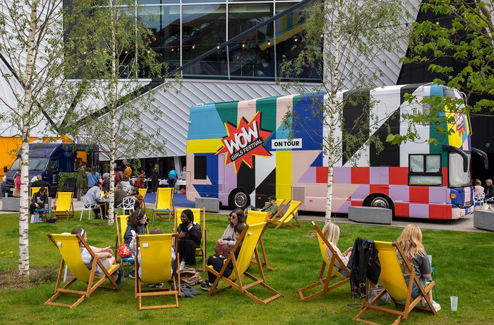 People relax on yellow deck chairs on a lawn, near a colourful bus with "WOW" graphic and a modern building in background