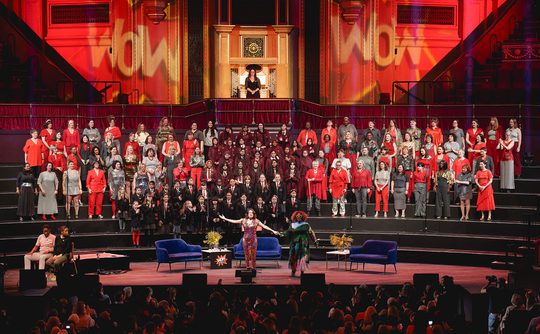 Concert in a grand hall with a large choir dressed in red, grey, and black. The lighting is vibrant and the audience is engaged
