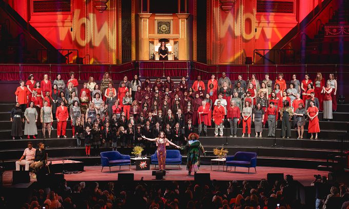 Concert in a grand hall with a large choir dressed in red, grey, and black. The lighting is vibrant and the audience is engaged