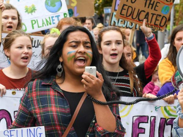 A diverse group of young protesters march, holding climate change signs. A girl with face paint speaks into a megaphone,
