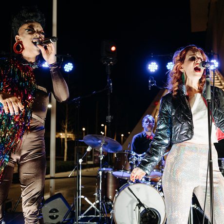 Two singers perform energetically on an outdoor stage at night, surrounded by band members.