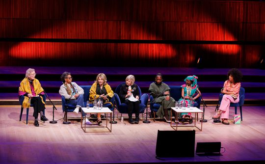 A diverse panel of seven people seated on stage, engaged in discussion. Warm lighting and wood paneling create an inviting atmosphere.