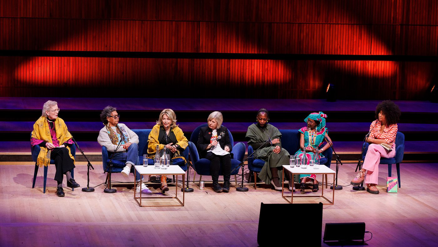 A diverse panel of seven people seated on stage, engaged in discussion. Warm lighting and wood paneling create an inviting atmosphere.