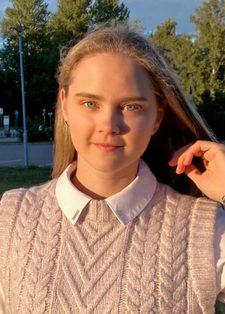 Veronika Podliesnova stands on some grass beside a car park, looking into the camera with her arm raised to her face. She has long blonde hair and is wearing a pink knitted tank top and white shirt.