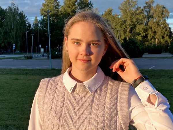 Veronika Podliesnova stands on some grass beside a car park, looking into the camera with her arm raised to her face. She has long blonde hair and is wearing a pink knitted tank top and white shirt.