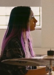 A young woman with long purple hair plays the drums in a sunlit room