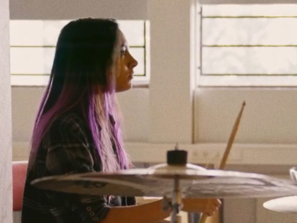 A young woman with long purple hair plays the drums in a sunlit room