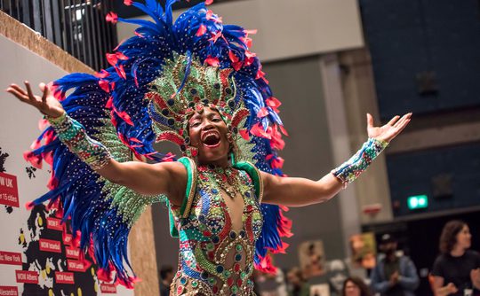 Dancer in vibrant, feathered costume joyfully performs with outstretched arms and smile
