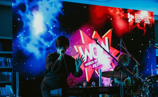 A musician performs on a dimly lit stage with a keyboard, set against a vibrant "WOW Women of the World Festival" backdrop in cosmic colours.