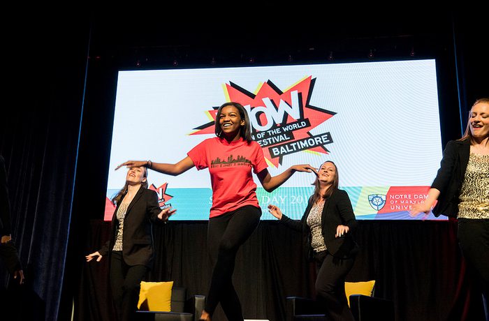 A woman in a red shirt energetically dances on stage, with three women in black outfits behind her. A "WOW Festival Baltimore" banner displays in the background.