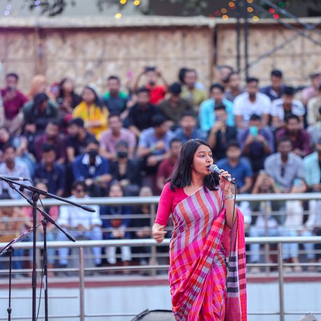 A woman in a colorful saree passionately sings into a microphone on stage. A lively crowd watches from bleachers, with bright stage lights nearby.