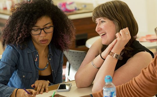 Two women sat at a table. One is concentrating hard and writing on a piece of paper, whilst the other is smiling and looking at something out of shot.