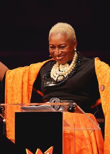 A person stands behind a podium with arms raised, wearing a vibrant orange shawl and a large necklace. They are smiling, conveying joy and confidence.