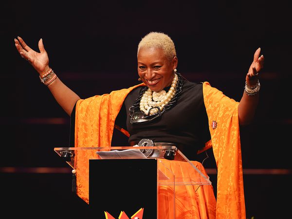 A person stands behind a podium with arms raised, wearing a vibrant orange shawl and a large necklace. They are smiling, conveying joy and confidence.