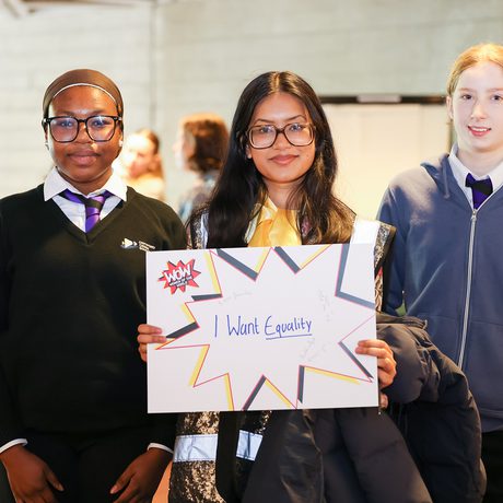 Three students stand together, smiling. The student in the center holds a sign that reads, "I Want Equality."