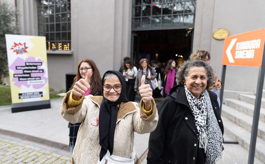 A group of smiling people exits a building, with a woman in front giving two thumbs up.