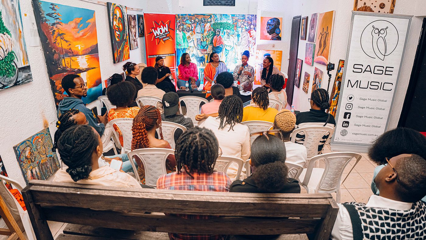 A group seated in a gallery listens to a panel discussion. Vibrant artwork fills the walls