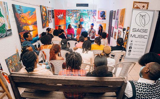 A group seated in a gallery listens to a panel discussion. Vibrant artwork fills the walls