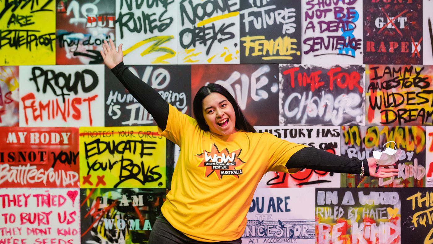 A person in a "WOW Australia" yellow shirt poses excitedly in front of a vibrant graffiti wall with empowering messages