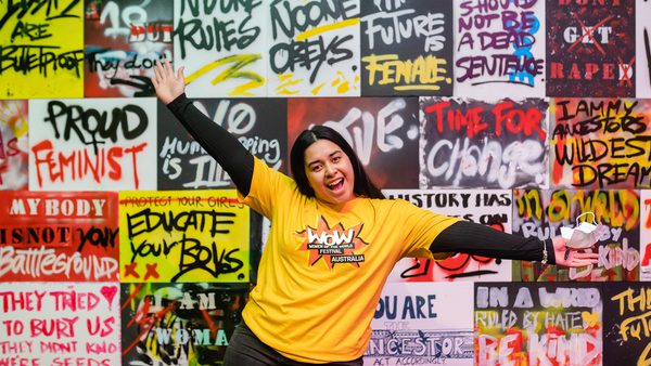 A person in a "WOW Australia" yellow shirt poses excitedly in front of a vibrant graffiti wall with empowering messages