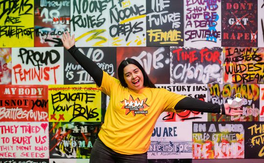 A person in a "WOW Australia" yellow shirt poses excitedly in front of a vibrant graffiti wall with empowering messages