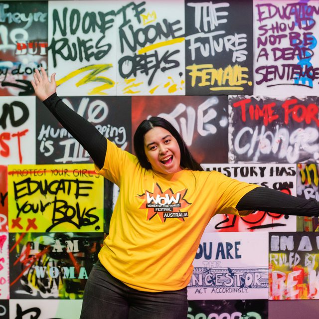 A person in a "WOW Australia" yellow shirt poses excitedly in front of a vibrant graffiti wall with empowering messages
