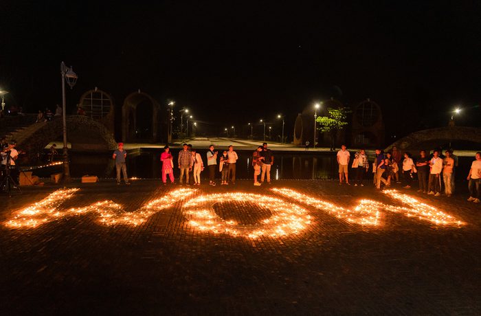 A group of people stand around candles arranged to spell "WOW" on the ground at night, creating a warm glow against the dark