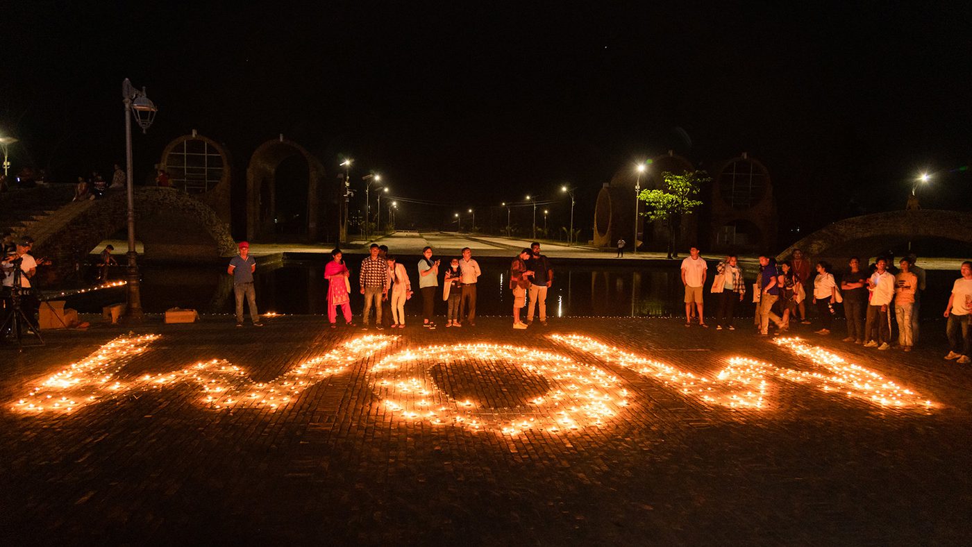 A group of people stand around candles arranged to spell "WOW" on the ground at night, creating a warm glow against the dark