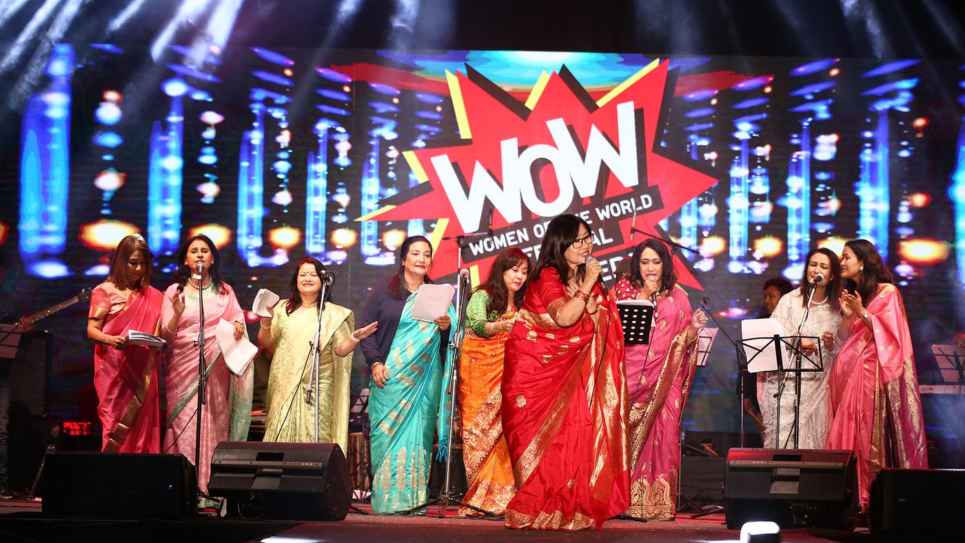 A group of women in colorful sarees on stage at the Women of the World event. Bright lights illuminate the scene, creating a joyful atmosphere.