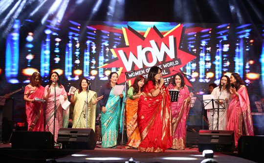 A group of women in colorful sarees on stage at the Women of the World event. Bright lights illuminate the scene, creating a joyful atmosphere.