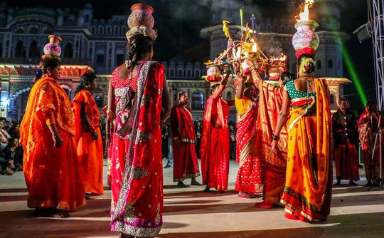 Women in vibrant red and orange sarees perform a dance at night, balancing decorated pots with flames on their heads.