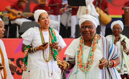 Elderly women in vibrant, traditional African clothing joyfully dance and play percussion instruments on stage with musicians in the background