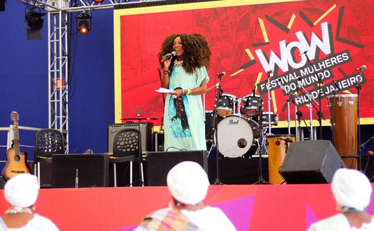 A woman speaks on stage at the WOW Festival Mulheres do Mundo in Rio. Bright decor, musical instruments, and an engaged audience set an inspiring scene.