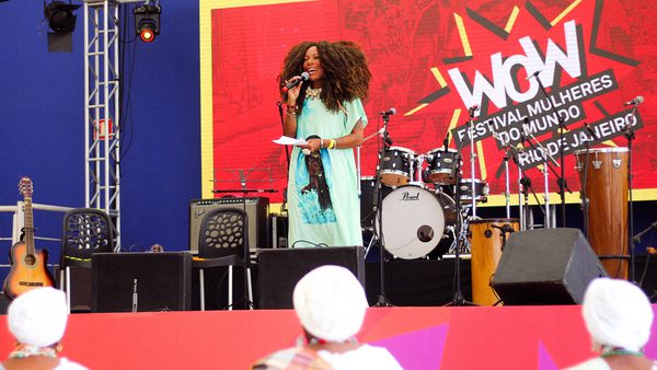 A woman speaks on stage at the WOW Festival Mulheres do Mundo in Rio. Bright decor, musical instruments, and an engaged audience set an inspiring scene.