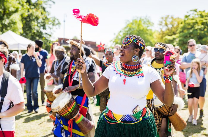 A joyful outdoor festival scene with a group wearing colourful attire, playing drums. The mood is vibrant and energetic, set against a sunny backdrop.