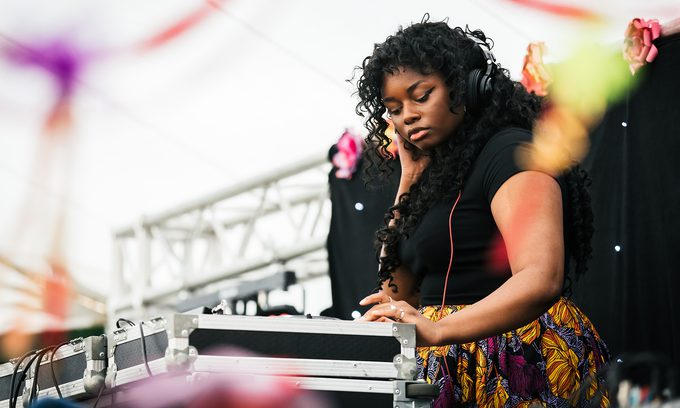Young DJ with curly hair focuses intently on mixing tracks at an outdoor event. She wears headphones and a colourful skirt.